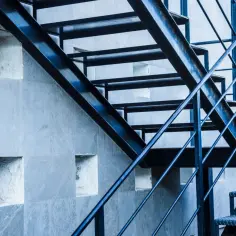 Metalowe schody z poręczą biegną wzdłuż szarej, kamiennej ściany z kwadratowymi wnękami. Murek zamiast balustrady tworzy ciekawy kontrast.