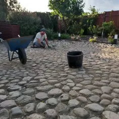 Mężczyzna pracuje przy budowie drogi z kamienia polnego. Obok stoi taczka i wiadro.