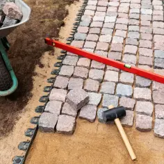 Układanie kostki brukowej na podbudowie z piasku, z użyciem poziomicy i gumowego młotka. W tle taczka z kamieniami.