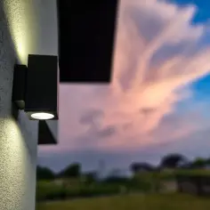 Nowoczesna lampa kinkietowa przymocowana do elewacji, oświetlająca ścianę. W tle zachód słońca z różowymi chmurami.