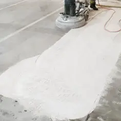 Maszyna szlifuje betonową posadzkę, tworząc biały pył. To przygotowanie do wypełnienia ubytków w posadzce betonowej.