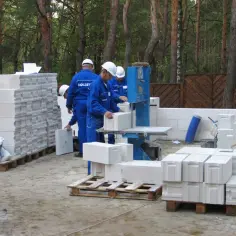 Budowa domu w lesie. Pracownicy w niebieskich kombinezonach układają bloczka betonowego na palecie.