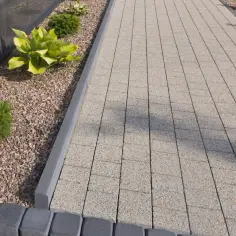 Nowy chodnik z kostki brukowej i obrzeży betonowych, otoczony kamyczkami i zielenią. Widać, ile betonu pod obrzeże zostało użyte do stworzenia solidnej podstawy.