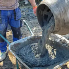 Robotnik w pomarańczowych rękawicach wylewa świeży beton z betoniarki do taczki. Zastanawiasz się, ile kosztuje tona betonu?