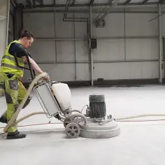 Pracownik w kamizelce odblaskowej szlifuje beton maszyną. To pokazuje, czym szlifować beton, by uzyskać gładką powierzchnię.