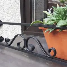 Elegancka barierka na parapet bez wiercenia, ozdobiona kutymi elementami, chroni doniczkę z zielonymi roślinami.