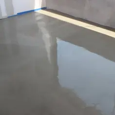 Świeżo wylany beton na posadzkę, błyszczący od wilgoci, czeka na stwardnienie. W tle widać ściany i niebieską taśmę.