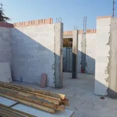 Budowa domu w toku. Widoczne betonowe słupy z wystającym zbrojeniem pod słup, ściany z bloczków gazobetonowych i drewno budowlane.