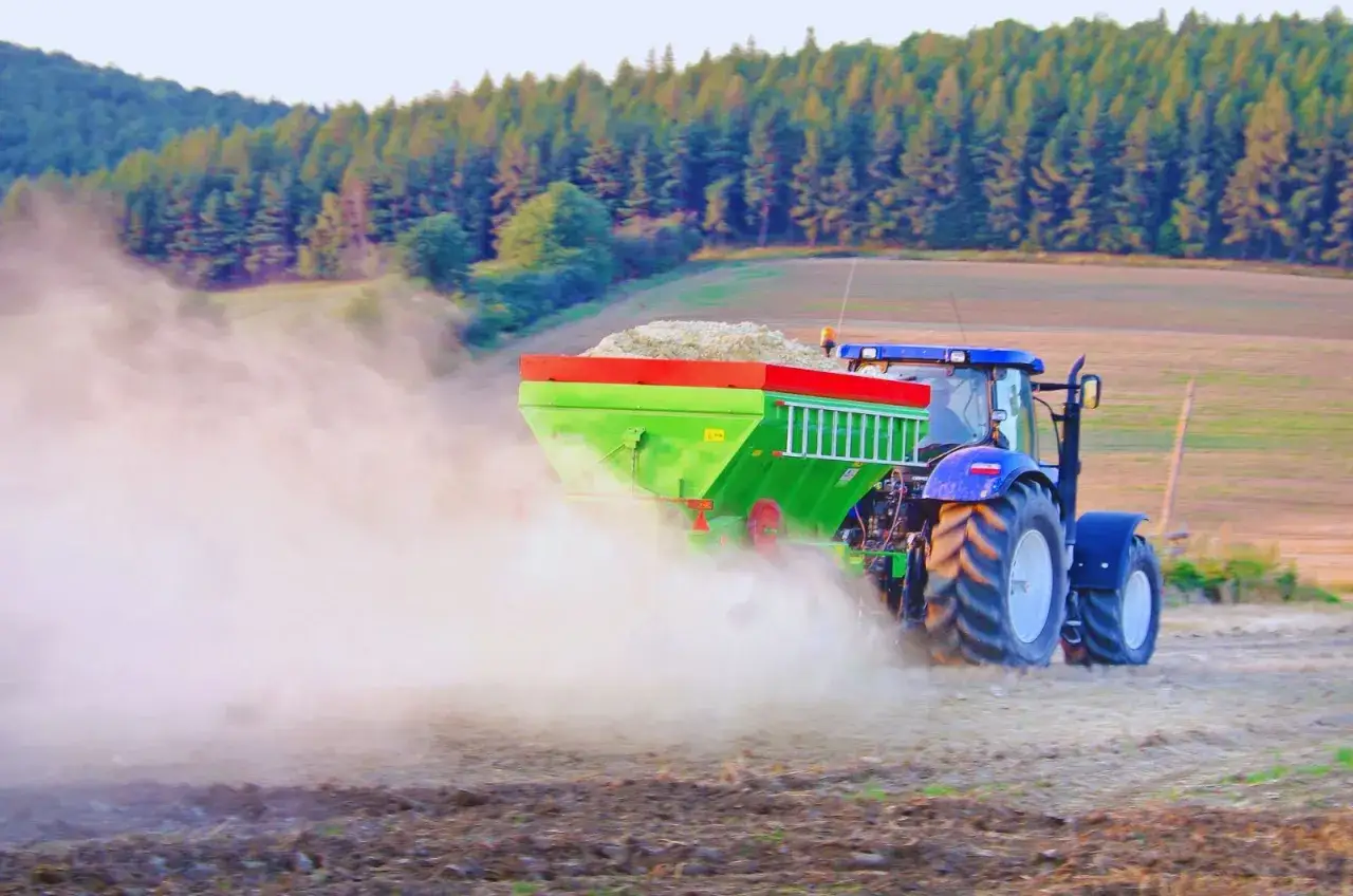 Traktor rozsiewa wapno węglanowo magnezowe na polu, tworząc chmurę pyłu.