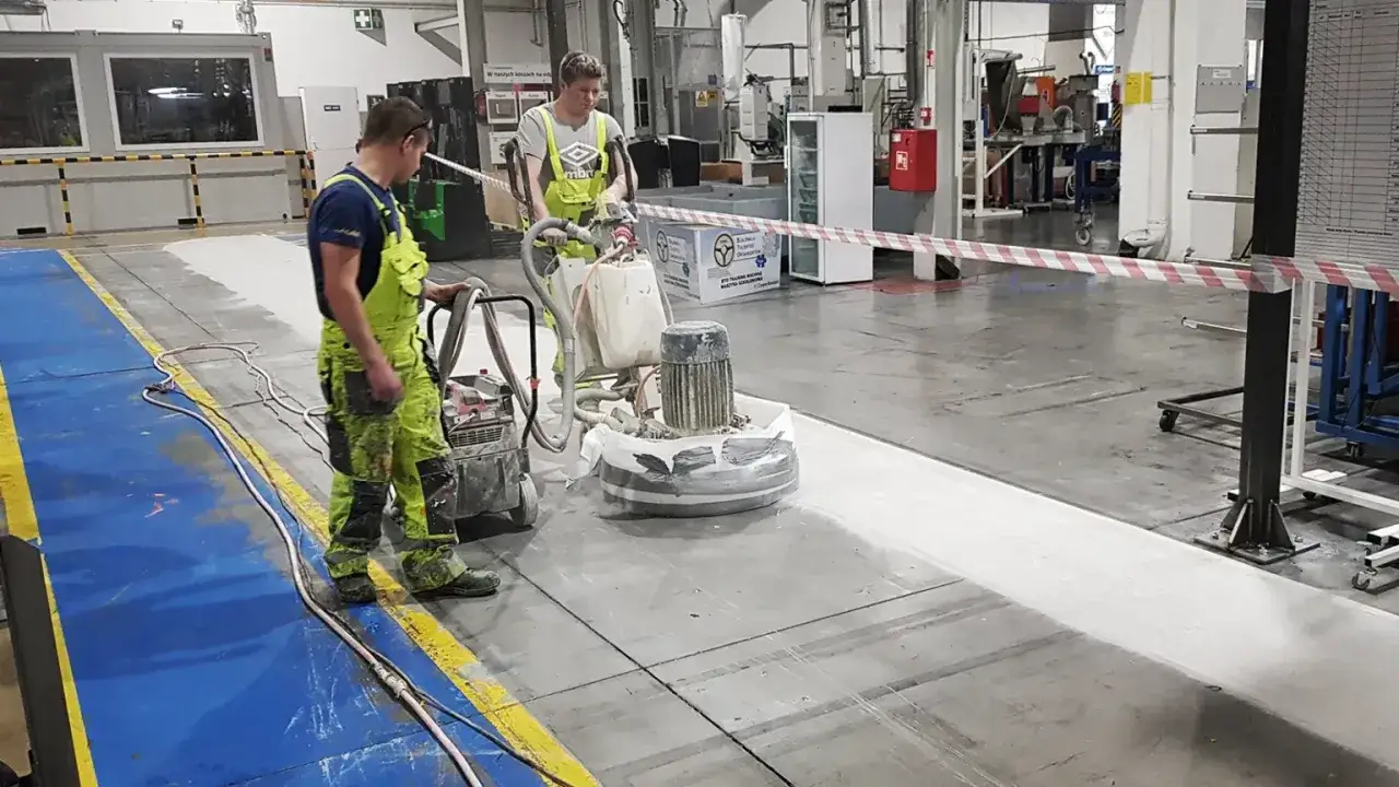 Dwóch pracowników w zielonych kombinezonach szlifuje betonową posadzkę przemysłową za pomocą specjalistycznej maszyny. To pokazuje, czym szlifować beton.
