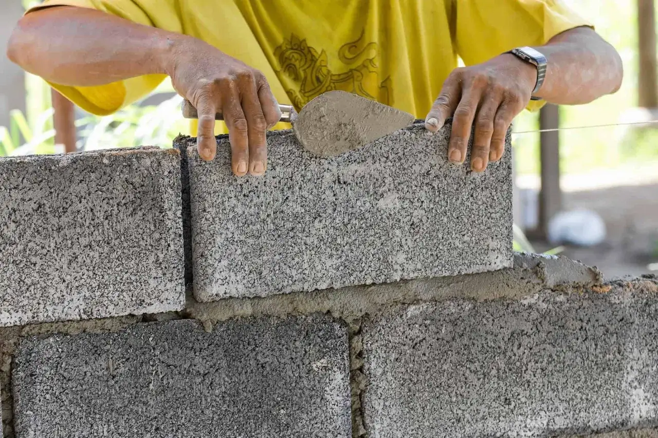 Ręce budowlańca układają kolejny bloczek betonowy na świeżej zaprawie, tworząc solidny mur.
