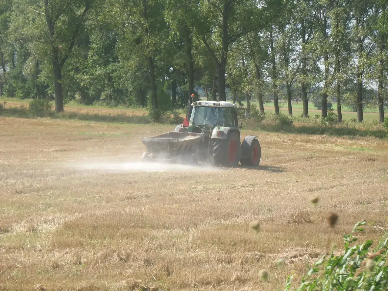Traktor rozsypuje wapno węglanowo magnezowe na polu, tworząc chmurę pyłu.
