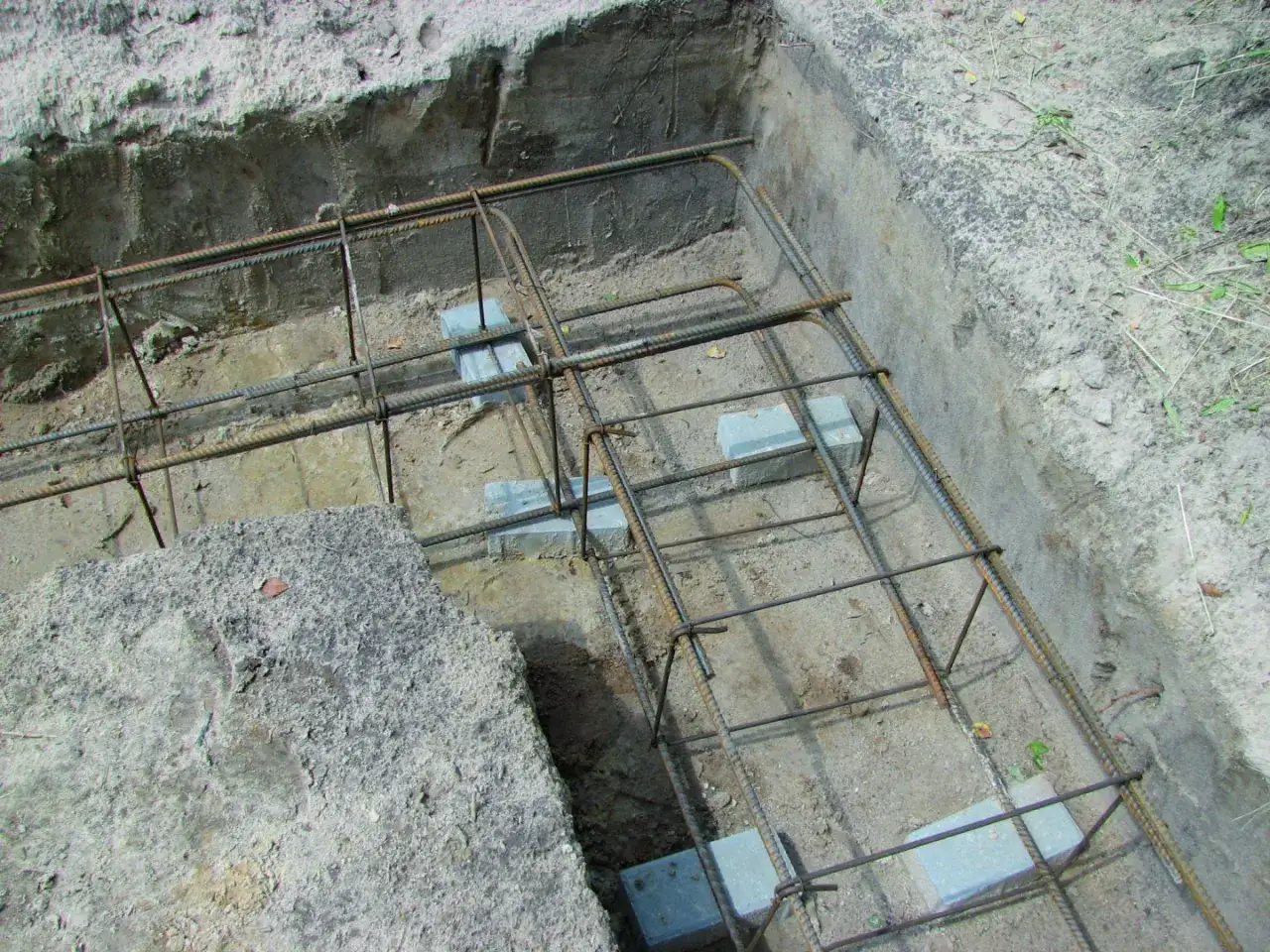 Gotowe zbrojenie ściany fundamentowej w wykopie. Stalowe pręty tworzą siatkę, podpartą na betonowych klockach, czekając na zalanie betonem.
