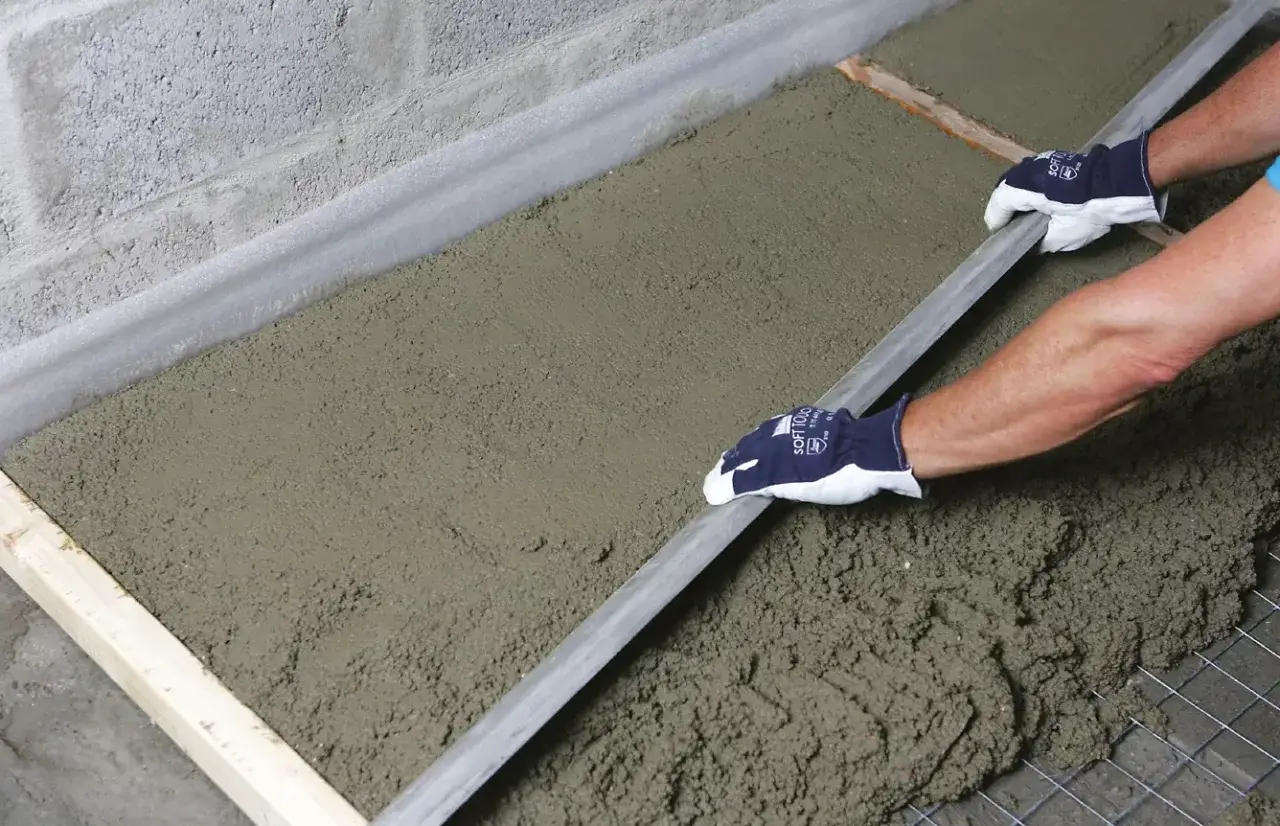 Ręce w rękawiczkach wygładzają świeży beton na wylewkę za pomocą łaty.