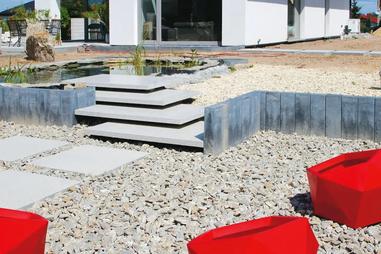 Nowoczesne schody z bloczków fundamentowych prowadzą do stawu. Otoczenie z kamieni i czerwone donice tworzą ciekawy ogród.
