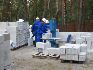 Budowa domu w lesie. Pracownicy w niebieskich kombinezonach układają bloczka betonowego na palecie.